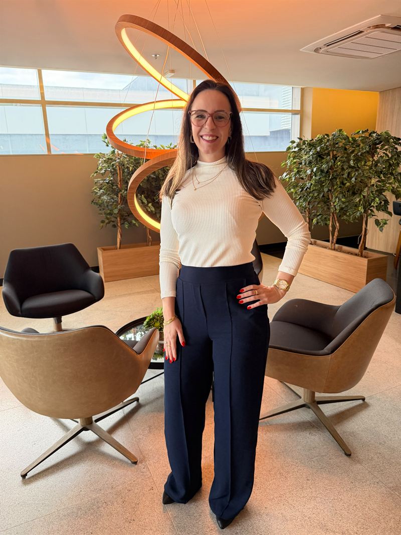 Carla Kerber em pé, sorrindo, num lobby com poltronas e plantas
