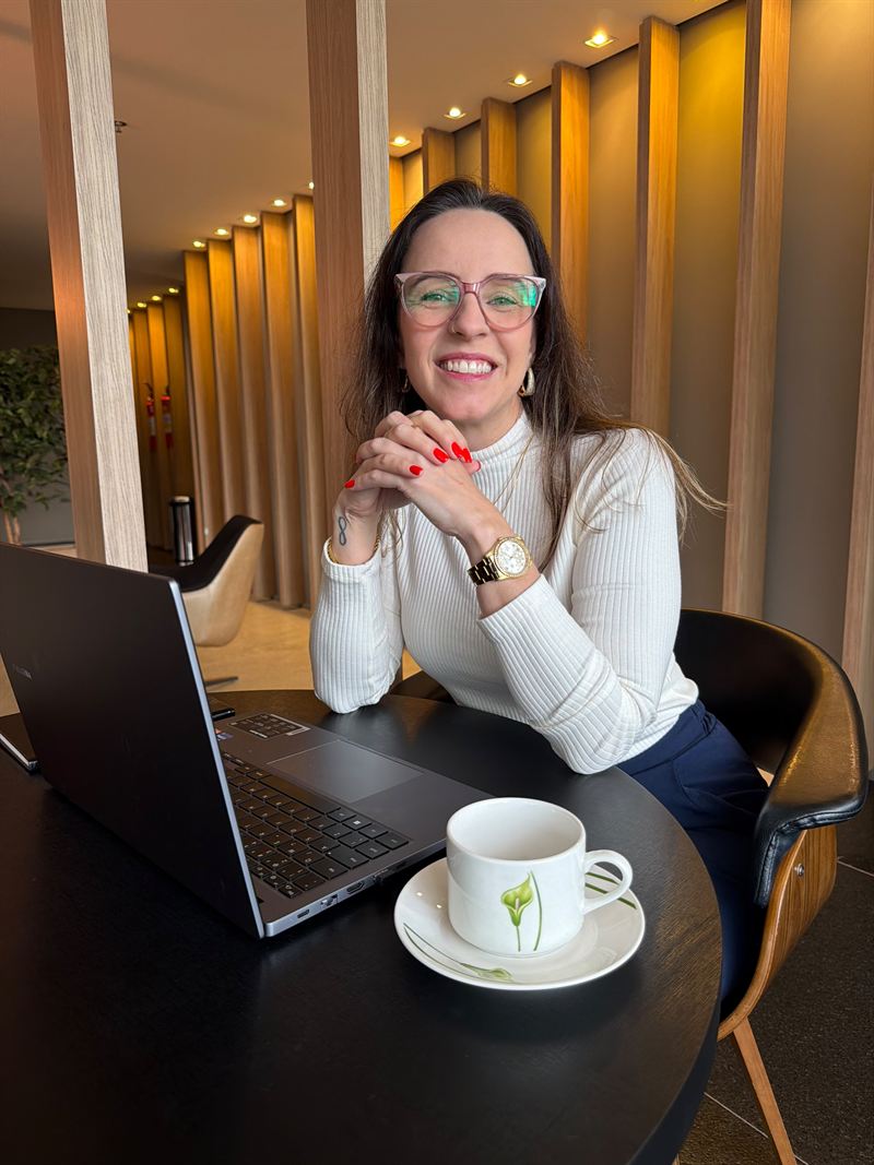 Carla Kerber sentada à mesa com o notebook aberto e uma xícara de café